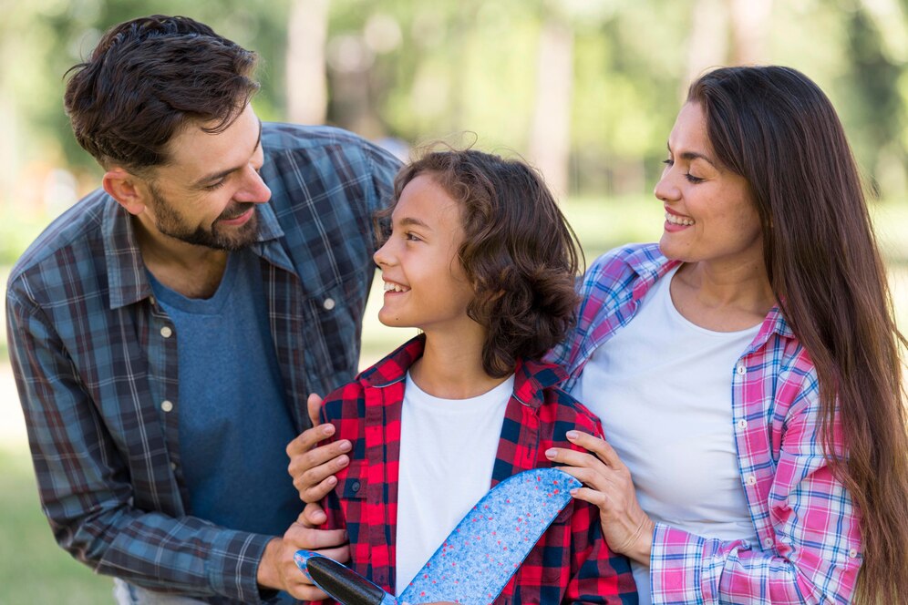 A family of three stands together in an outdoor setting, wearing casual clothing, with a lush green background. The child holds a colorful object.