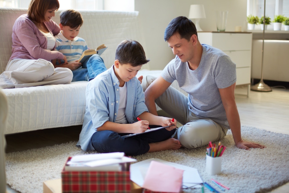 A family enjoys a cozy indoor moment; a child writes while sitting on a rug, and others relax on a sofa reading and engaging.