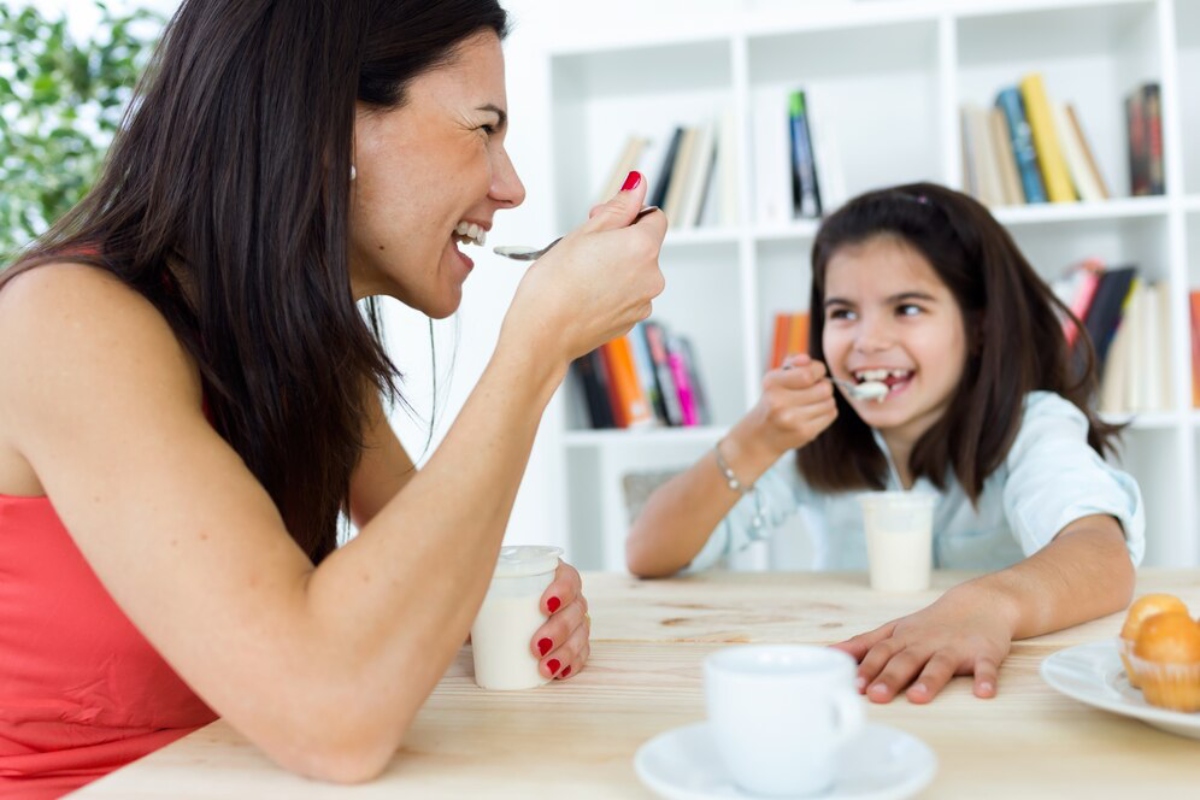 A woman and a child enjoy yogurt together at a wooden table, with a plate of pastries and a cup nearby, surrounded by books.