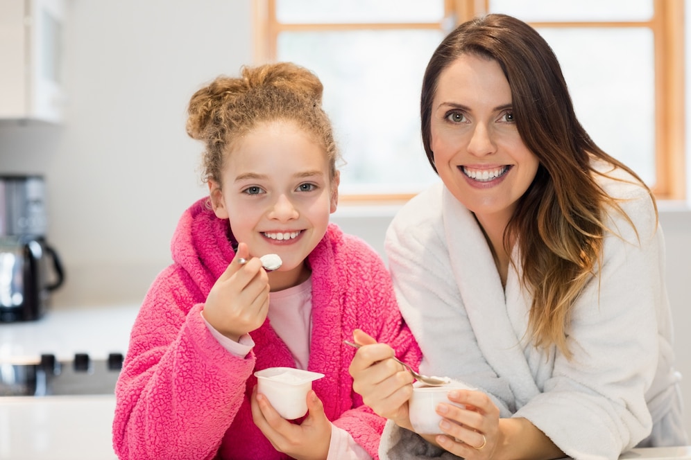 A woman and a girl in cozy bathrobes enjoying yogurt together at a kitchen table with natural light streaming through the window.