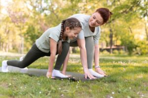 A young girl and an adult woman are practicing yoga stretches on a mat in a sunny park, surrounded by lush greenery.