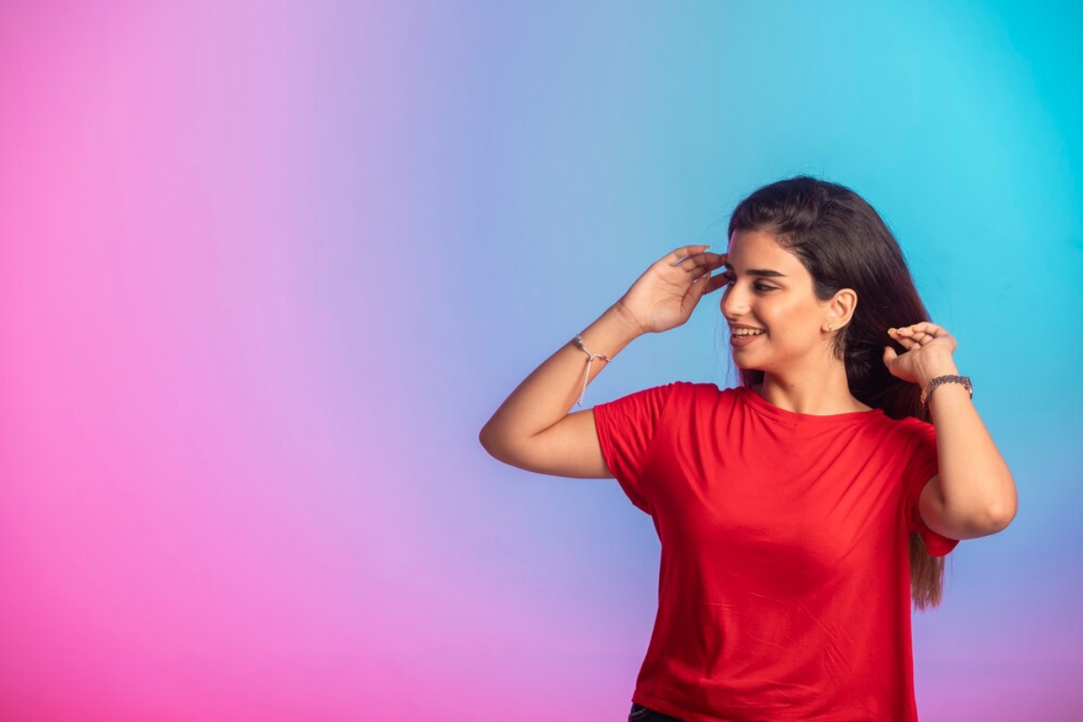 A person wearing a red t-shirt adjusts their hair against a vibrant pink and blue gradient background.