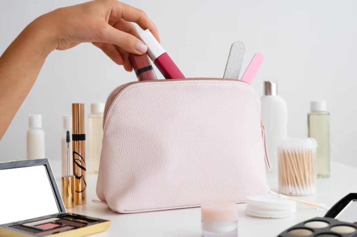 A hand placing makeup products into a pink cosmetic bag, surrounded by skincare and beauty items on a white table.