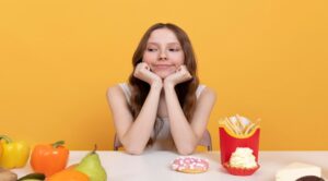 A person sits at a table covered with colorful fruits and various snacks, including fries, ice cream, and a donut, against a bright yellow background.