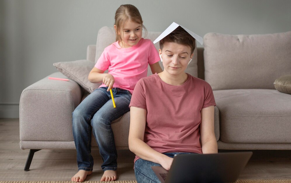 A child playfully balances a paper on an adult's head while sitting on a couch, with a laptop in front of the adult.