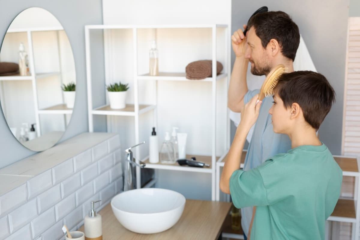 A parent and child stand together in a modern bathroom, brushing hair in front of a circular mirror and wooden sink.