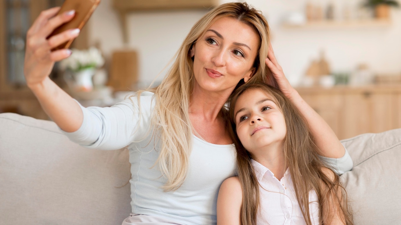 A woman and a young girl sit closely together on a sofa, taking a selfie with a smartphone in a cozy, warmly lit room.