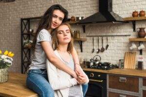 Two young women share a cozy moment in a stylish kitchen, with wooden counter, modern appliances, and a vase of flowers nearby.
