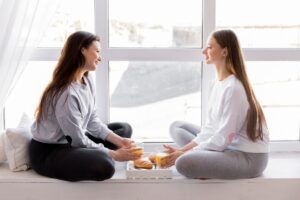 Two women sit cross-legged by a bright window, sharing drinks and pastries on a tray, surrounded by soft pillows.