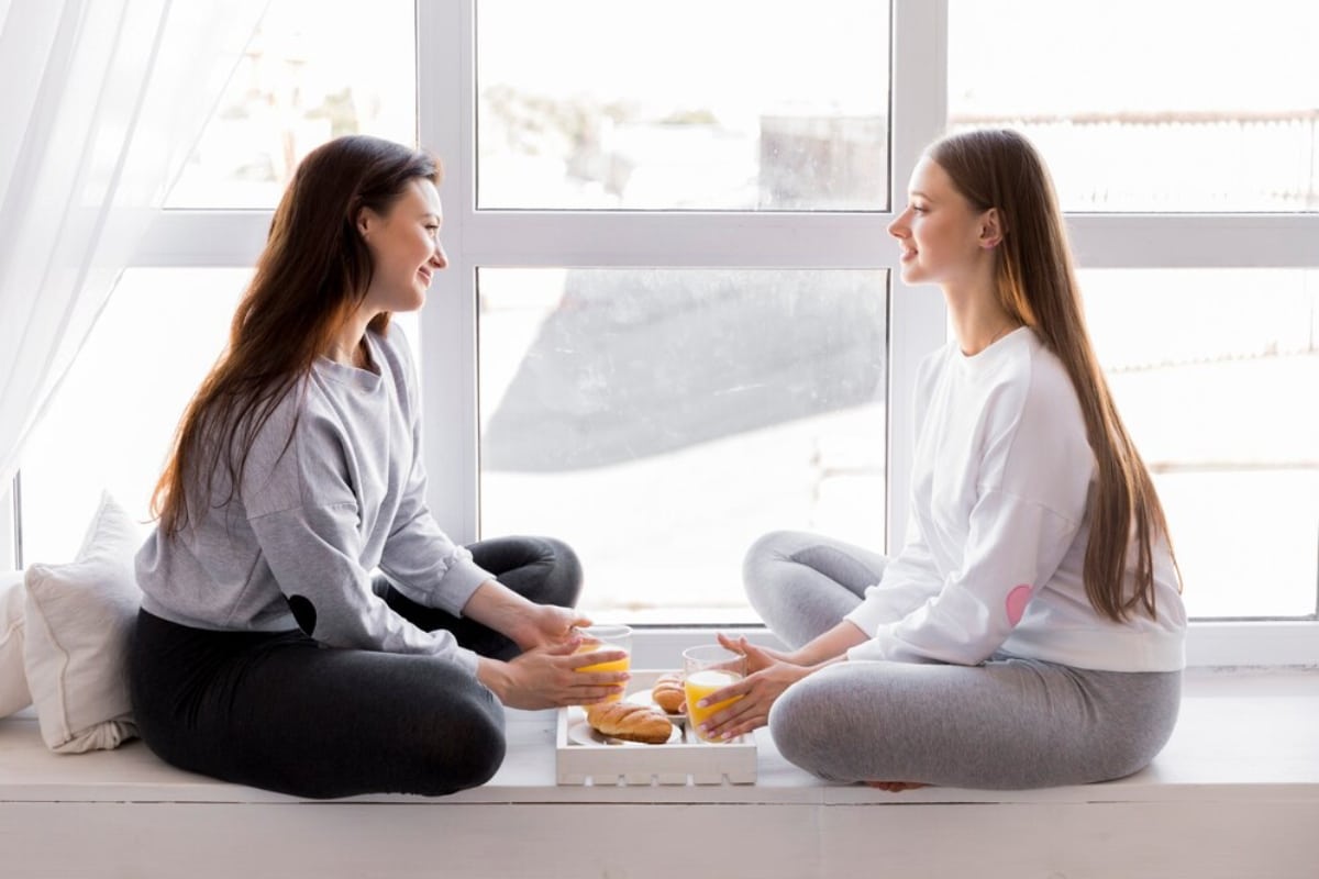 Two women sit cross-legged by a bright window, sharing drinks and pastries on a tray, surrounded by soft pillows.