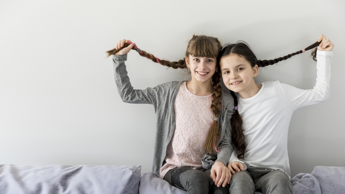 Two children sitting on a sofa, playfully holding each other's braided hair against a plain white wall.