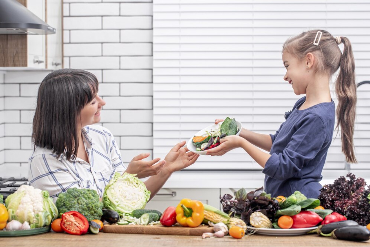 A child hands a plate of fresh vegetables to an adult in a bright kitchen surrounded by a variety of colorful produce.