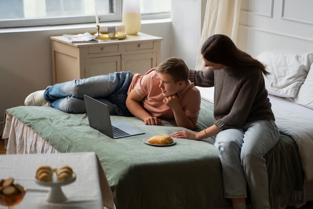 A woman tenderly assists a child on a bed with a laptop, while a plate of food sits nearby in a cozy, sunlit room.