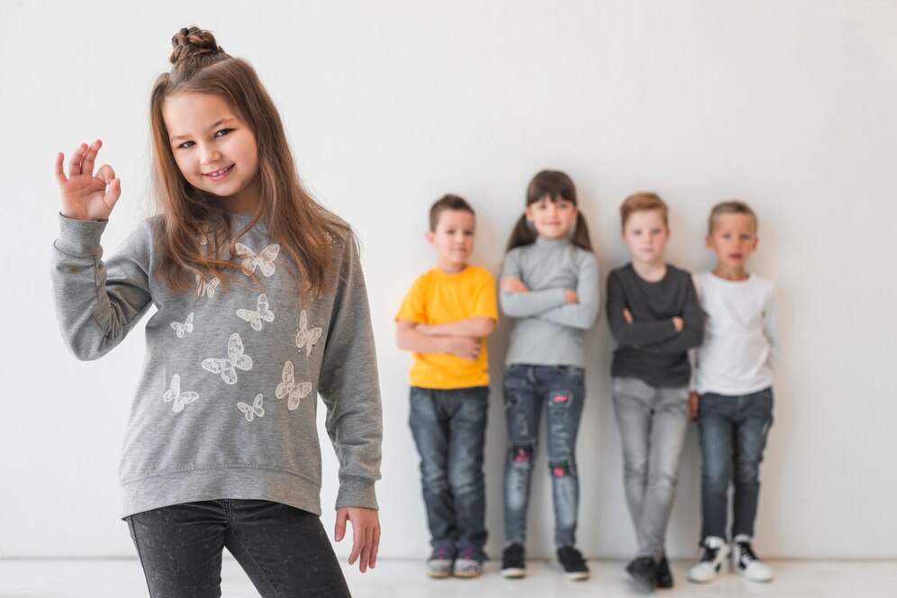 A girl in a butterfly-patterned sweatshirt shows an OK gesture, with four other children standing casually behind her.