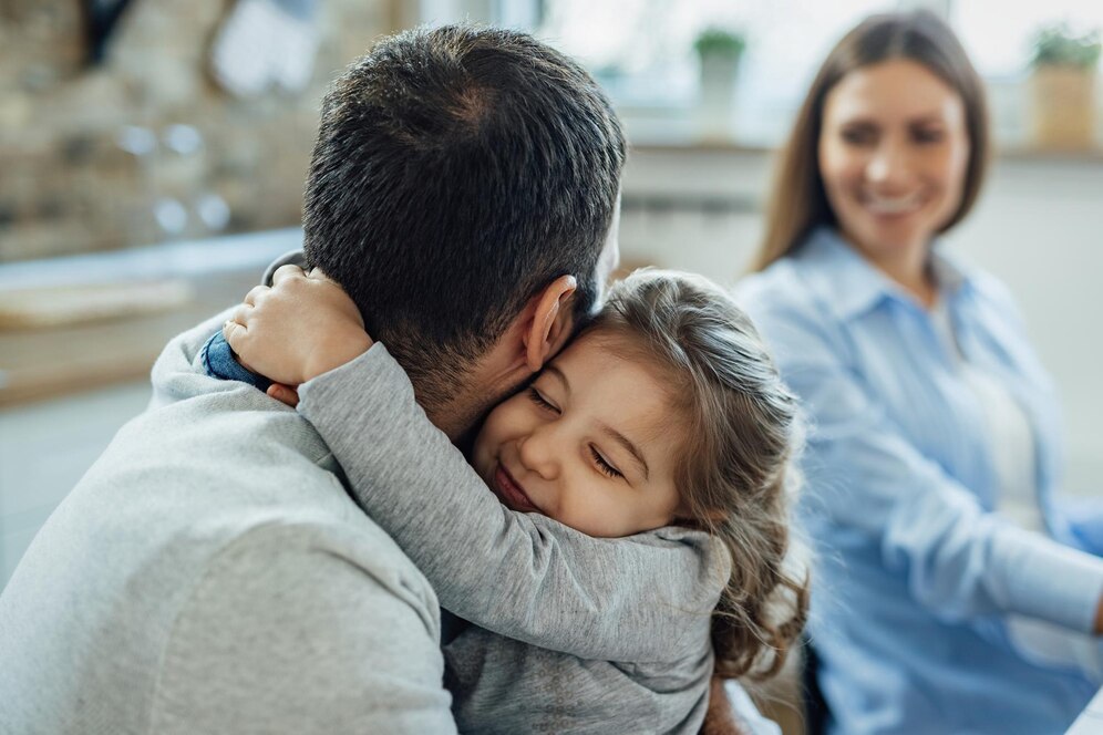 A child hugs an adult from behind in a cozy, softly lit room, with a third person seated nearby, all conveying a warm, intimate atmosphere.