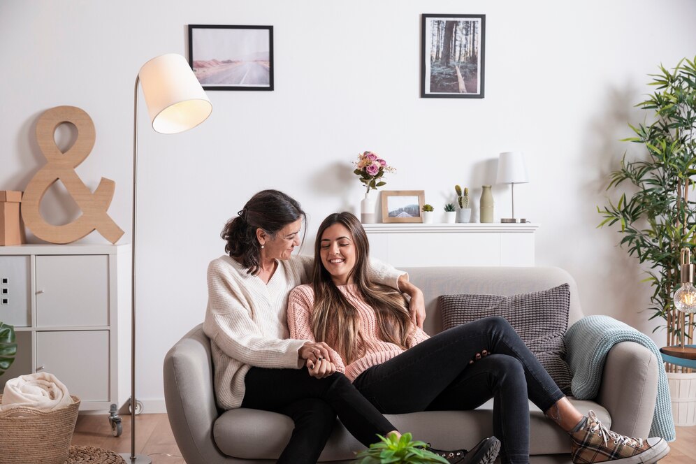 Two friends sit close together on a light gray couch in a cozy living room, decorated with plants and artwork on the walls.