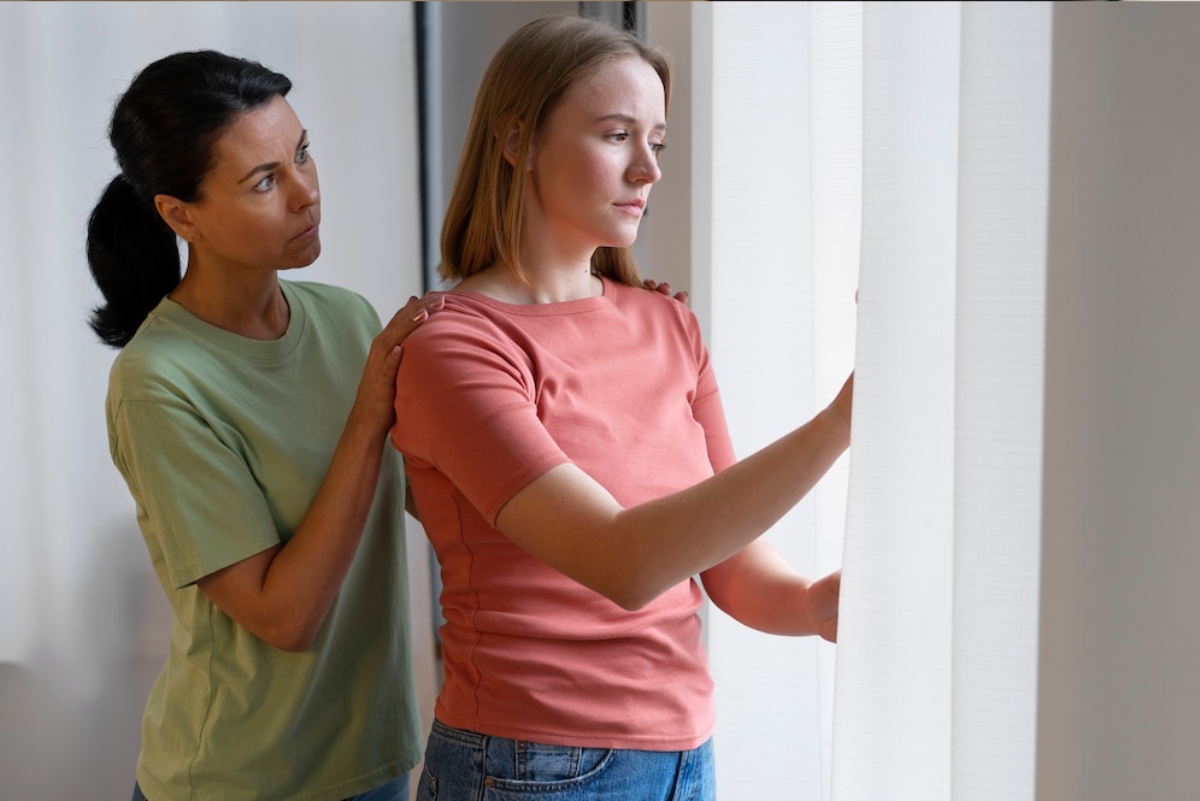 Two women stand indoors near a window, one gently touching the shoulder of the other, who is looking out at the light.
