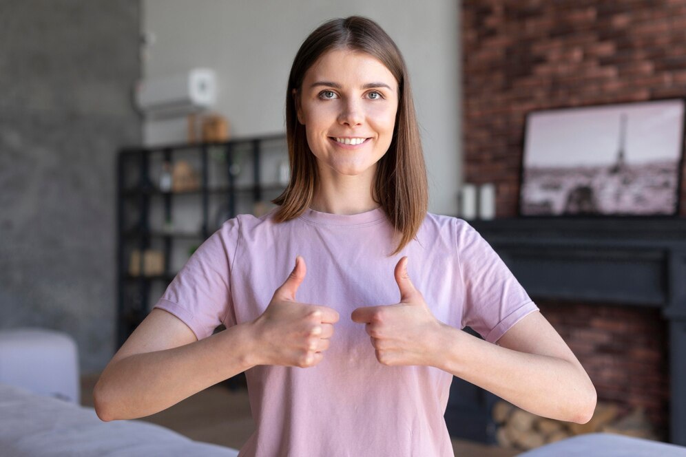 A person in a pink t-shirt is giving two thumbs up, smiling confidently in a cozy indoor setting with a modern aesthetic.