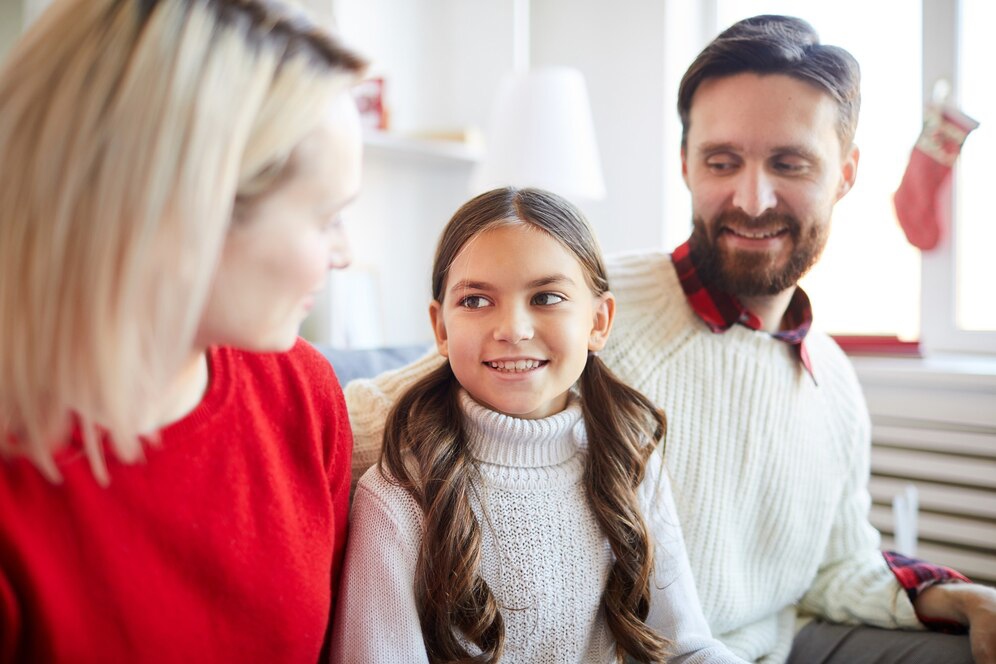 A family of three sits together indoors, dressed in cozy sweaters, enjoying a warm, festive atmosphere. Natural light brightens the room.