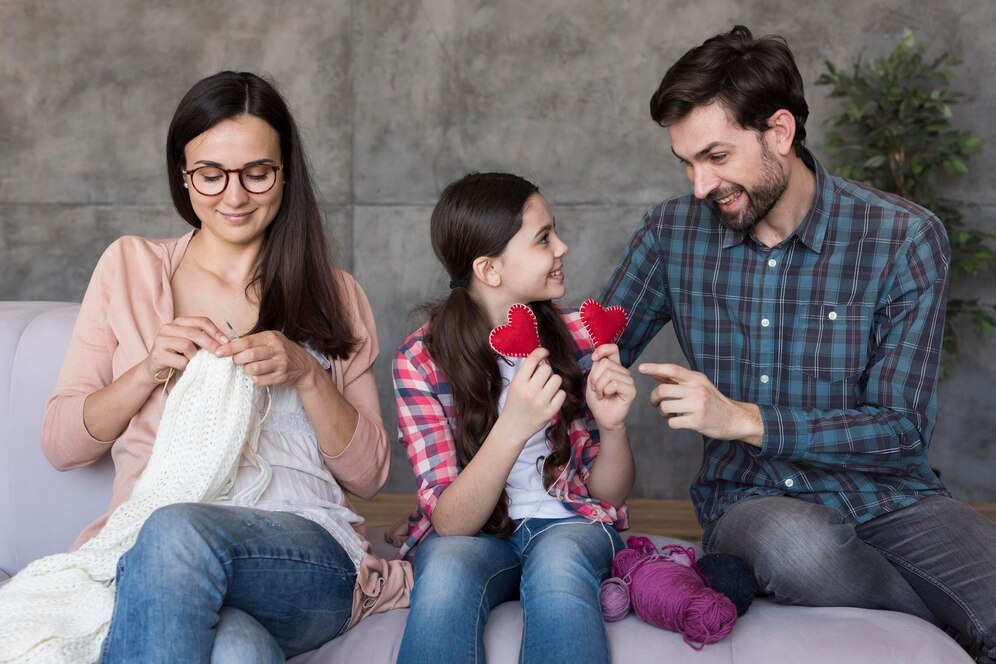 A woman knits while a girl holds red heart-shaped crafts, with a smiling man engaging with them, all seated comfortably.