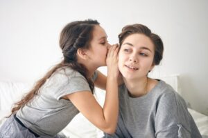 A child whispers a secret to an adult in a cozy, softly lit room, both dressed in casual gray outfits.