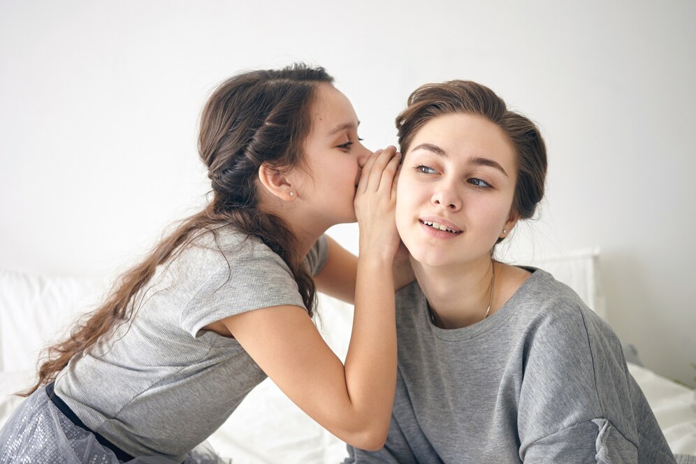 A child whispers a secret to an adult in a cozy, softly lit room, both dressed in casual gray outfits.