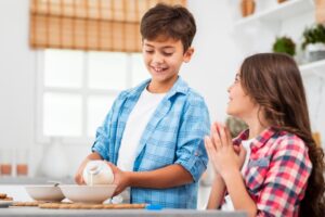 Two children are in a bright kitchen, one pouring milk into a bowl while the other watches eagerly, hands clasped in excitement.