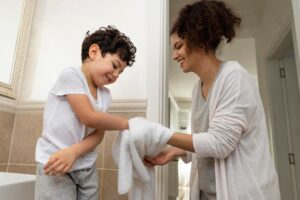 A child in a white T-shirt receives a towel from a woman in a cozy sweater, both engaging in a caring bathroom routine.