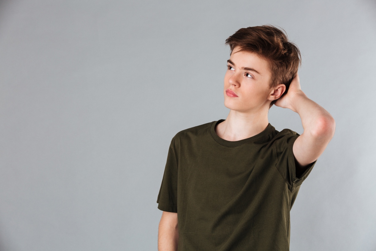 A young man in a dark green t-shirt poses against a light gray background, one hand behind his head and a thoughtful expression.