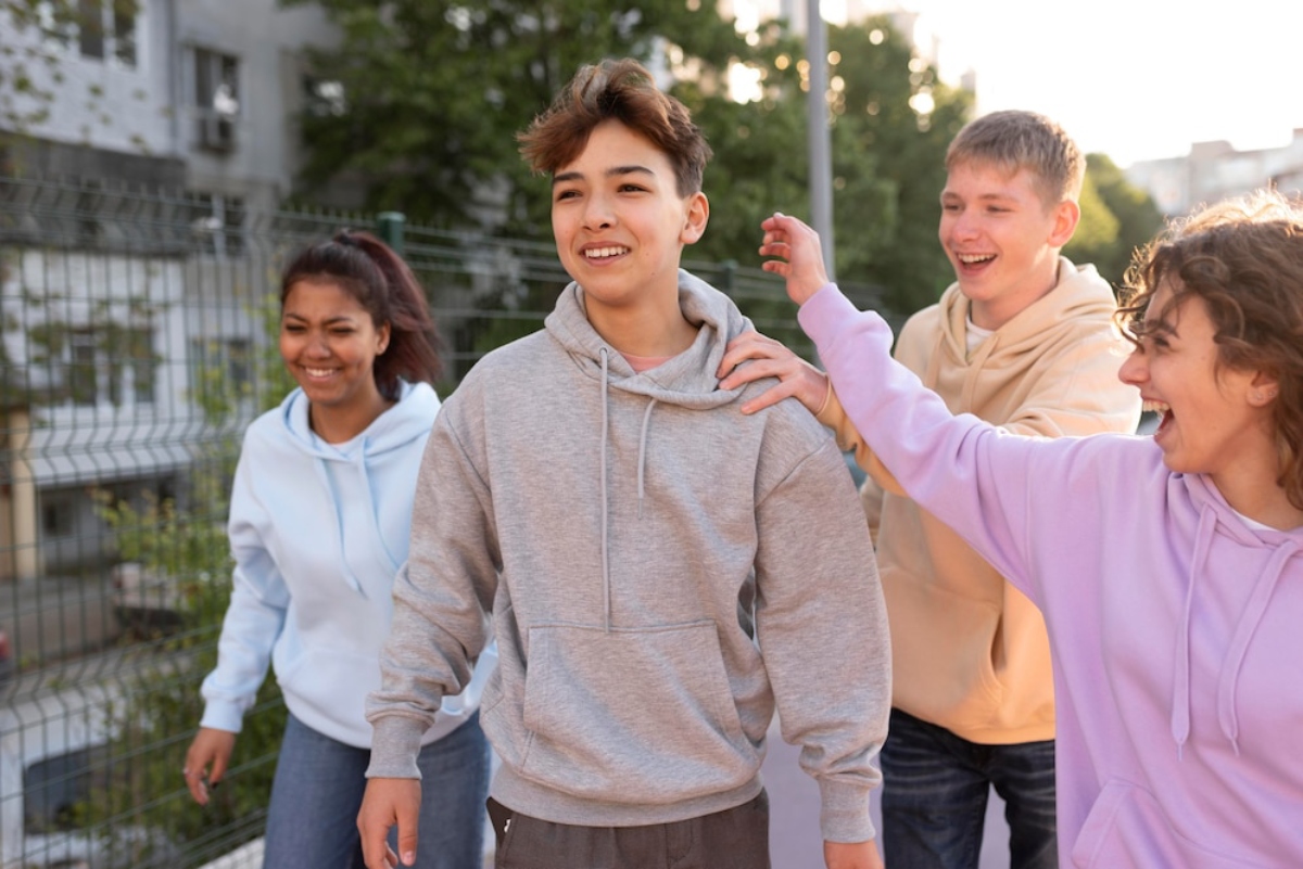 A group of young people in hoodies interact joyfully outdoors, surrounded by greenery and urban scenery.
