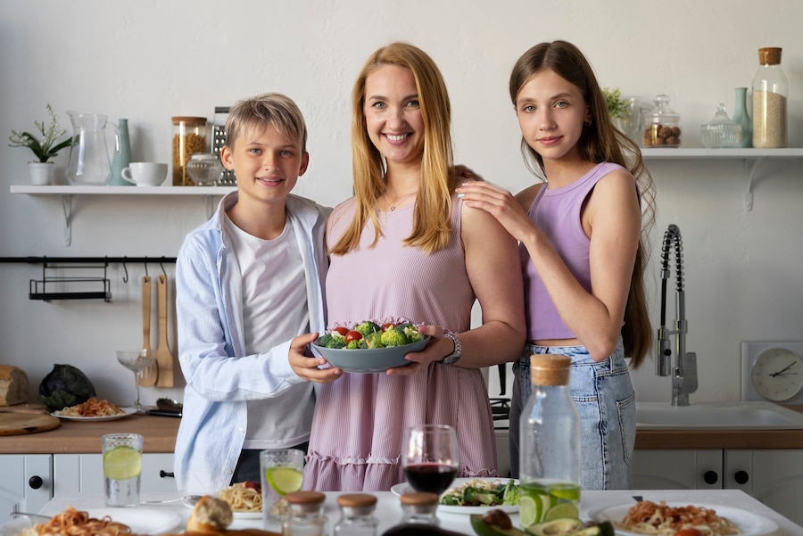 A group of three individuals in a modern kitchen, posing with a vibrant salad bowl, surrounded by various dishes and drinks on the table.