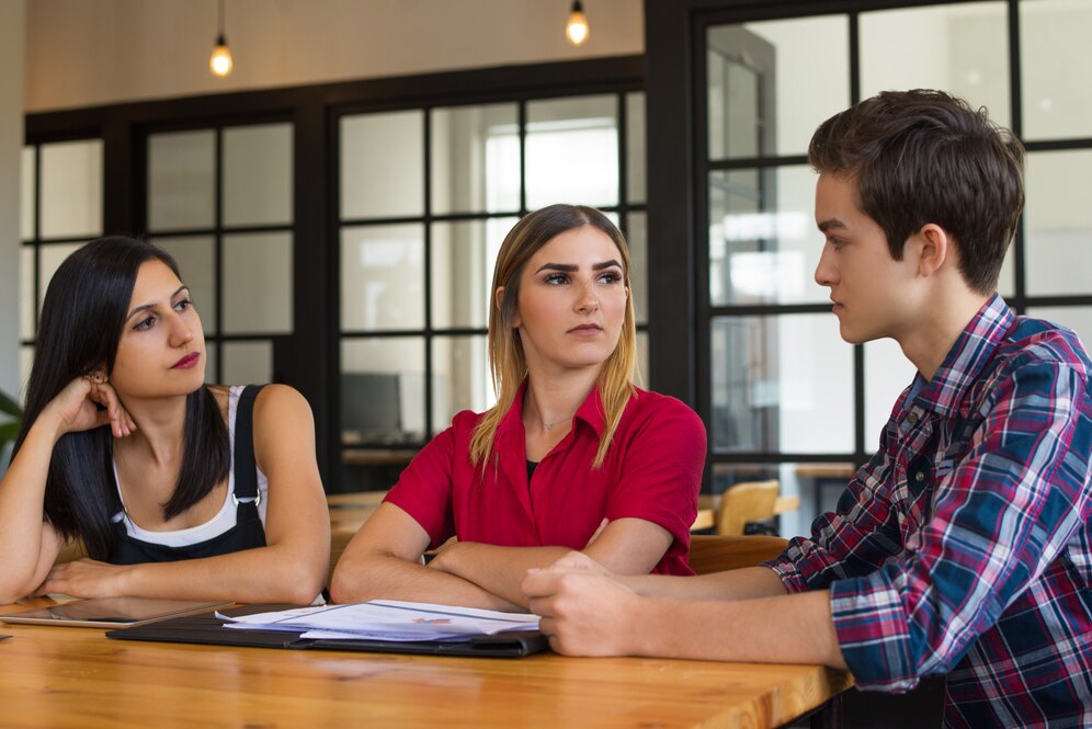 Three young adults engage in a serious conversation at a wooden table in a modern, bright café. Papers are spread out in front of them.