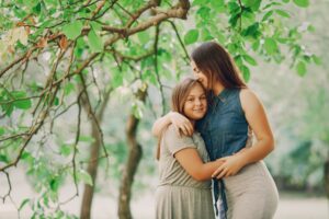 Two girls embrace warmly under a leafy tree, surrounded by lush greenery, depicting a moment of friendship and closeness.
