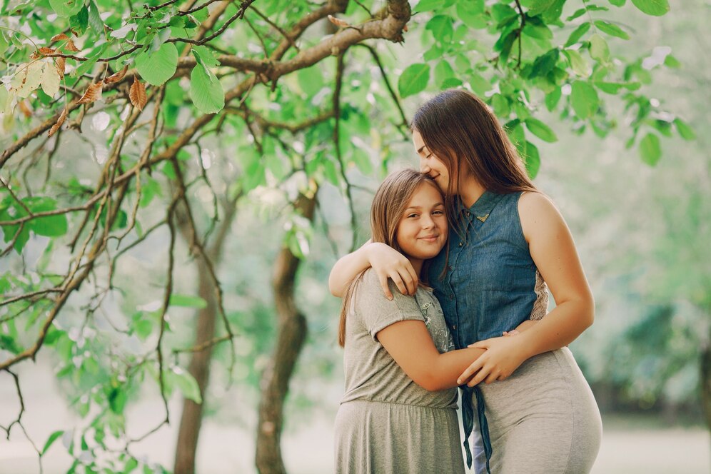 Two girls embrace warmly under a leafy tree, surrounded by lush greenery, depicting a moment of friendship and closeness.