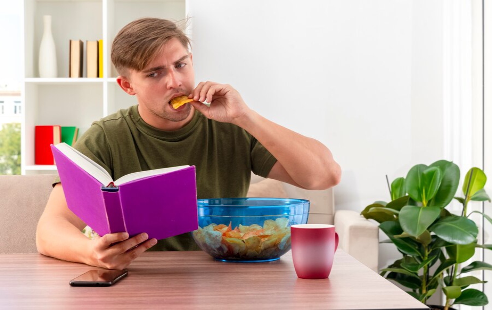A person sits at a wooden table, reading a purple book while snacking from a blue bowl of chips, with a red cup nearby and plants in view.