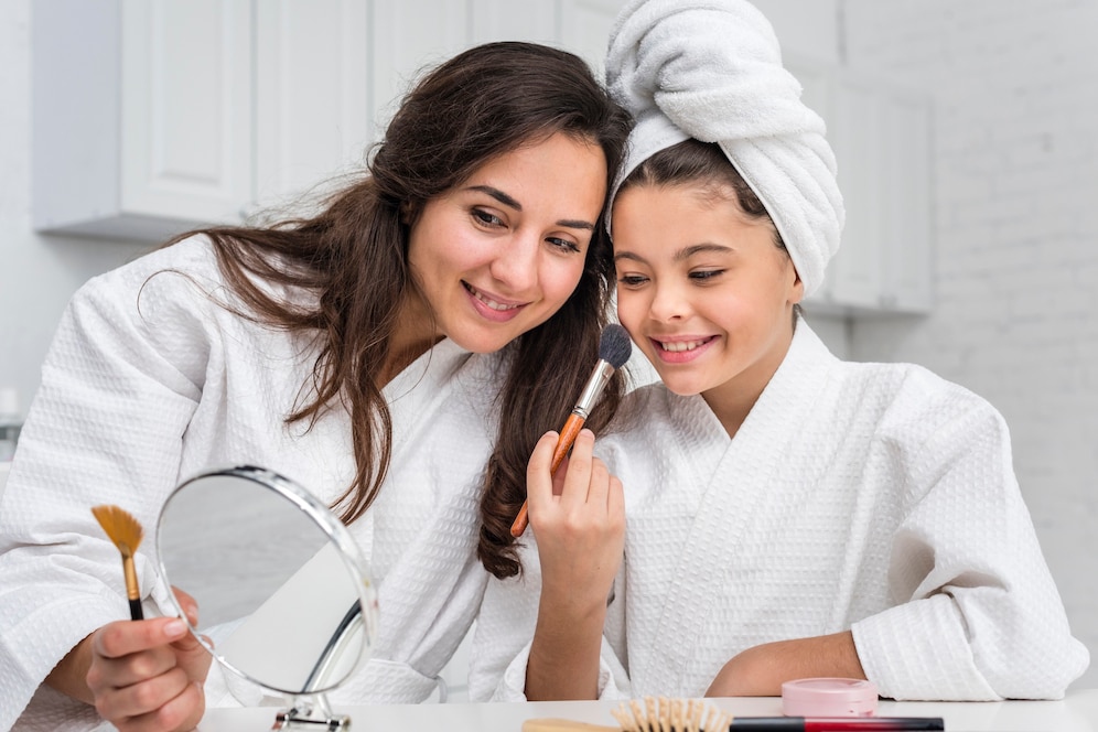 A woman and a child, both in towels, apply skincare products in a cozy bathroom setting, sharing a joyful skincare moment together.