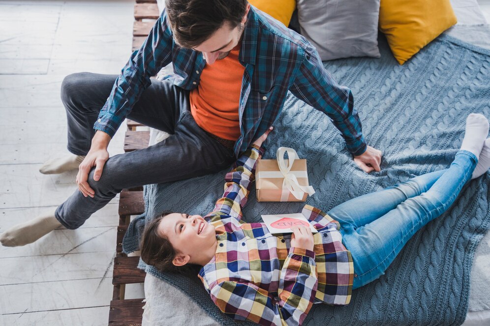 A young couple sits on a cozy bed, with a gift box and a handwritten card placed between them, surrounded by colorful pillows.
