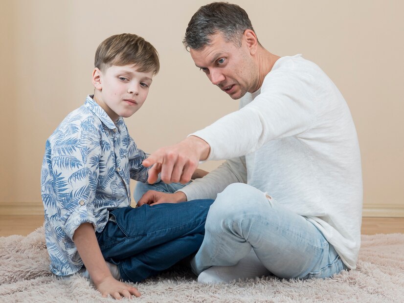 A child in a blue floral shirt sits cross-legged while an adult gestures in conversation, both on a cozy rug in a minimalistic room.