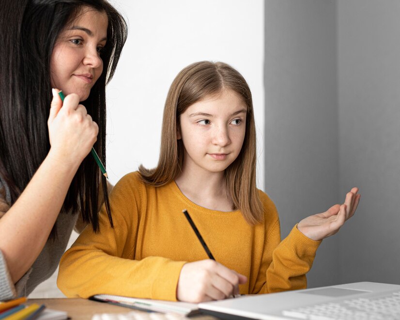 Two individuals engage in a discussion, one gesturing while the other writes notes at a desk with a laptop and colored pencils.