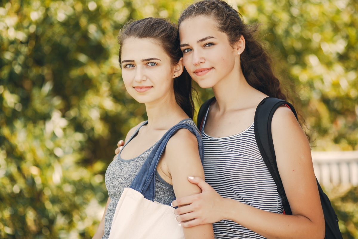 Two teenage girls standing together outside, smiling at the camera with arms around each other, wearing casual outfits and backpacks.
