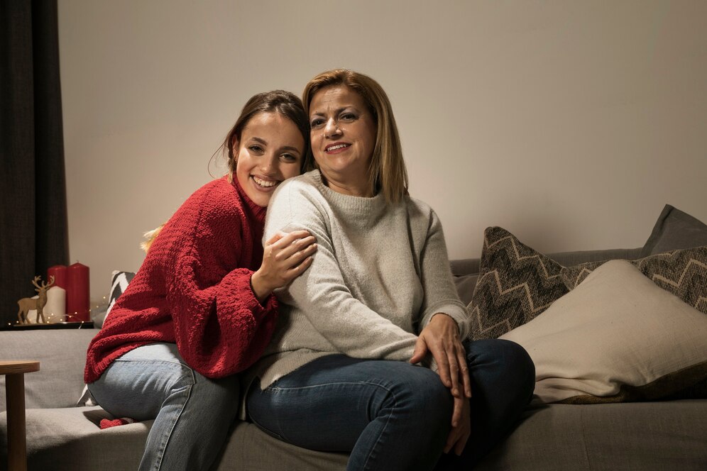 A woman in a cozy gray sweater sits on a couch, while a girl in a red sweater leans affectionately against her shoulder.