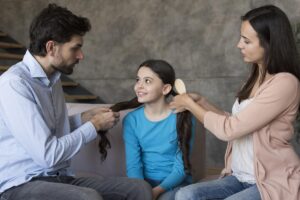 A child sits between two adults, one weaving hair into a braid while the other gently brushes it, set in a cozy, modern interior.