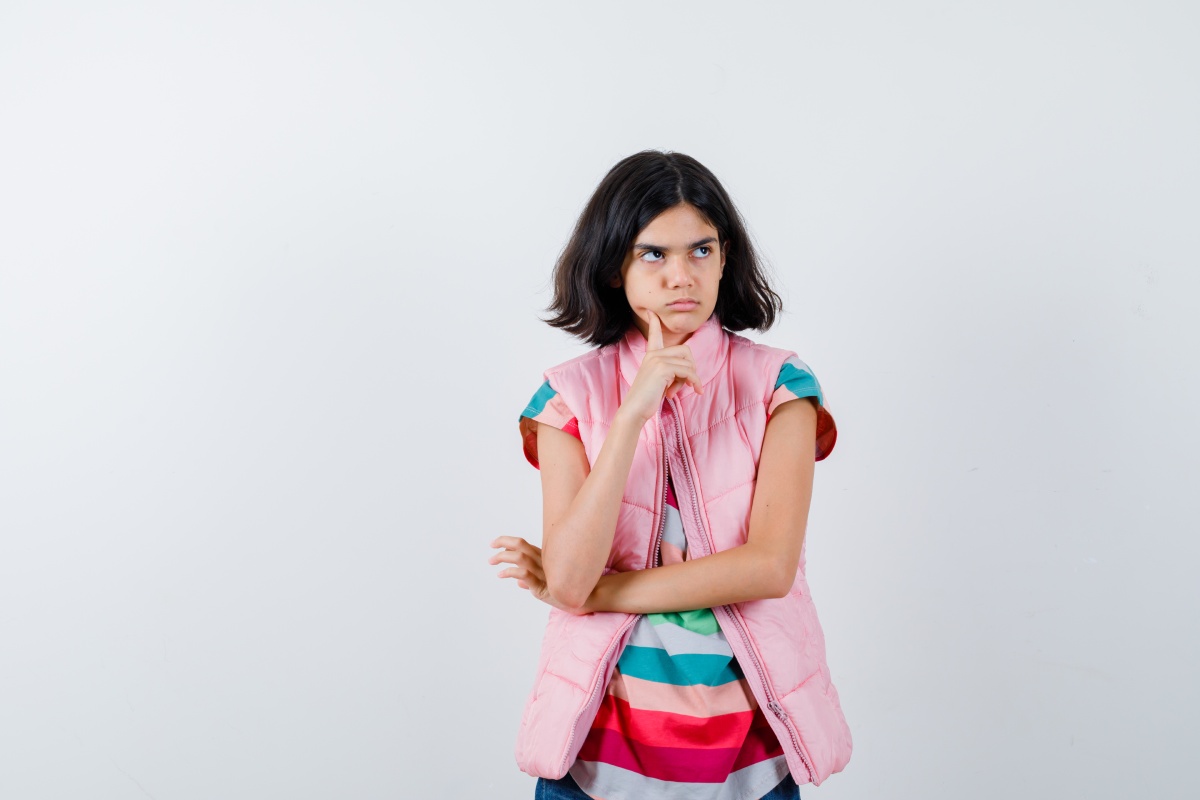 A young girl in a colorful striped shirt and pink vest standing against a white background, looking thoughtful with her hand on her chin.