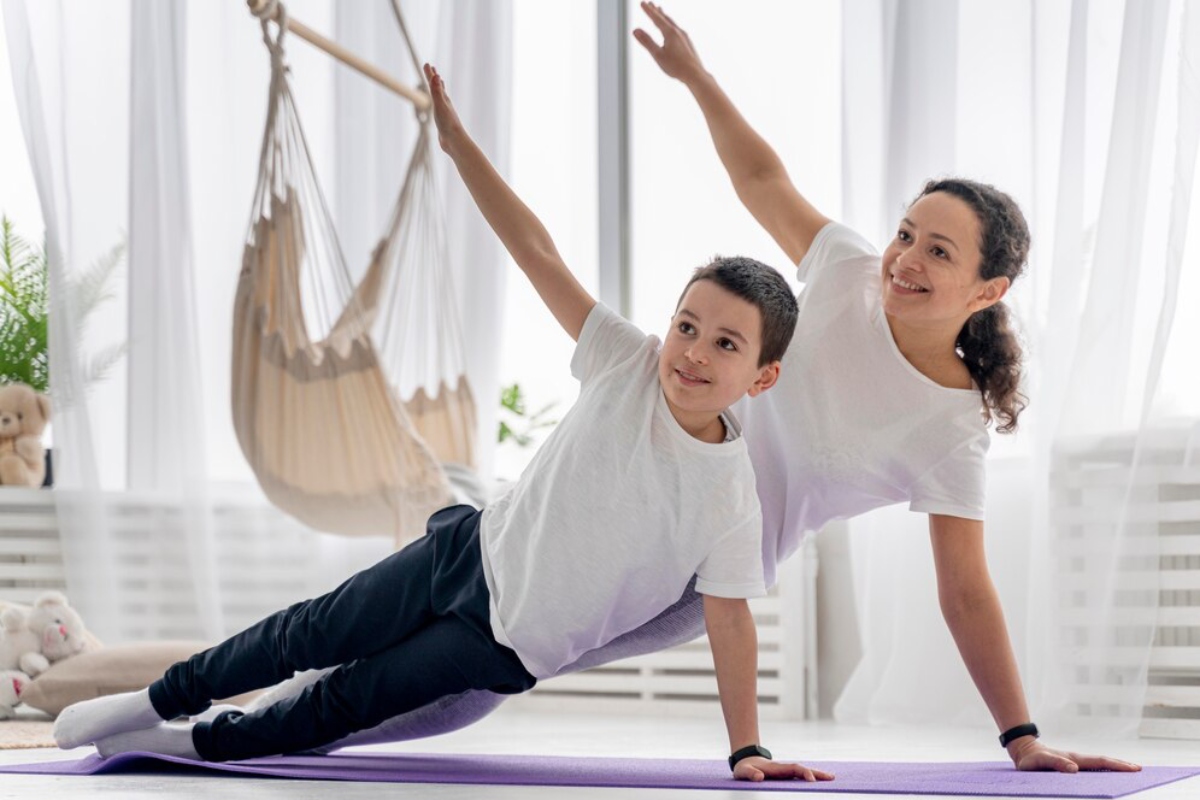 A child and an adult do a side plank exercise on a purple yoga mat in a bright, airy room adorned with plants and a hanging chair.