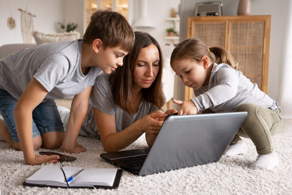 A woman and two children focus intently on a laptop while seated on a cozy, light-colored carpet with notepad and pen nearby.
