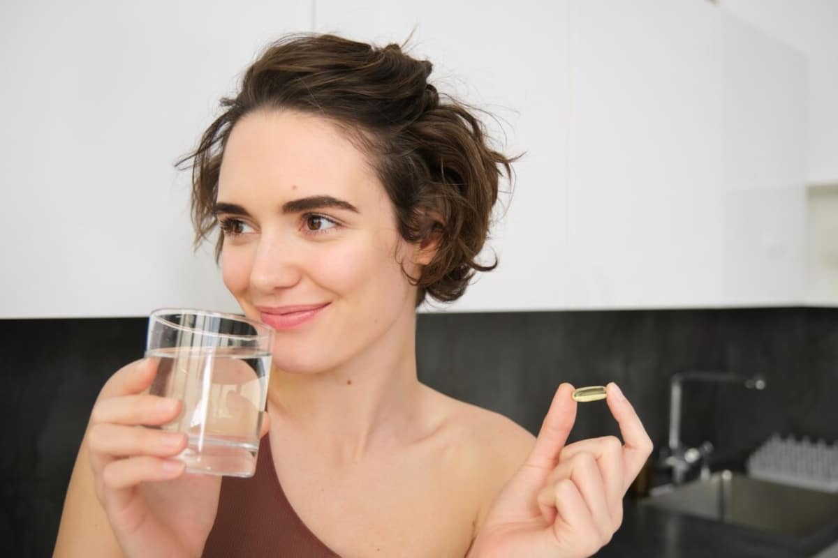 A person holding a glass of water in one hand and a yellow supplement capsule in the other, set in a modern kitchen.